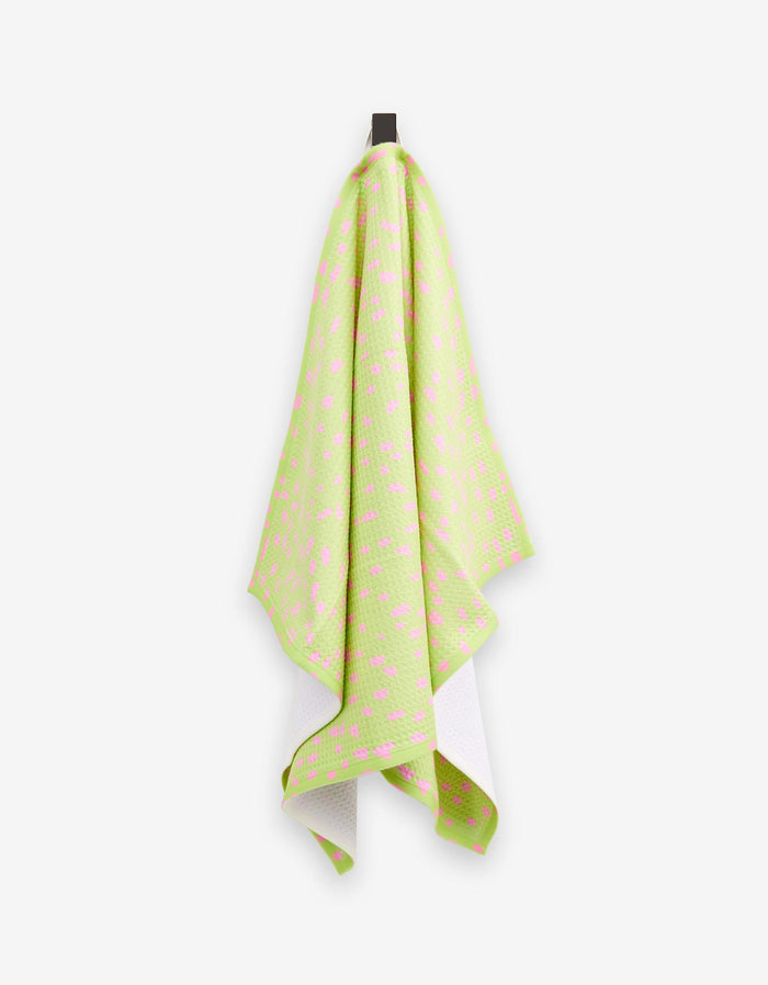 The GEOMETRY Speckle Green tea towel, featuring a pink geometric pattern and soft Geoweave fabric, hangs on a hook against a white background. Its slightly curved edges add a modern accent to your kitchen.