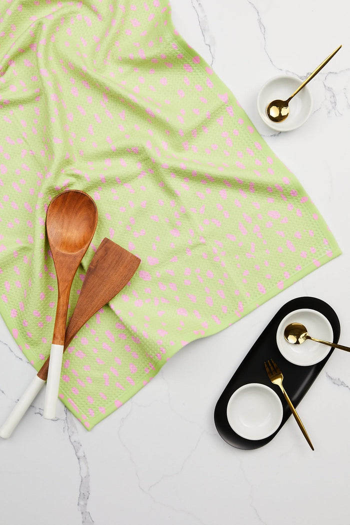 The Speckle Green tea towel by GEOMETRY, made from soft, durable Geoweave fabric, is paired with two wooden utensils with white handles and three small white bowls with gold spoons on a white marble surface.