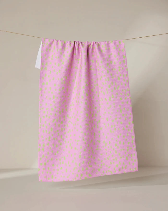 The GEOMETRY Speckle Pink tea towel, featuring ultra-absorbent fabric and small irregular green spots, hangs draped over a clothesline against a plain beige background.