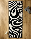 The GEOMETRY Spiral yoga mat features bold black and white abstract waves, shown on a light wooden floor with a recycled yoga towel. A water bottle and two gray blocks complete the setup.
