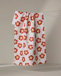 The Spotty Floral tea towel by Linda Fox, crafted from recycled materials, features a white background with red and yellow flowers and hangs neatly over a thin line against a neutral backdrop.