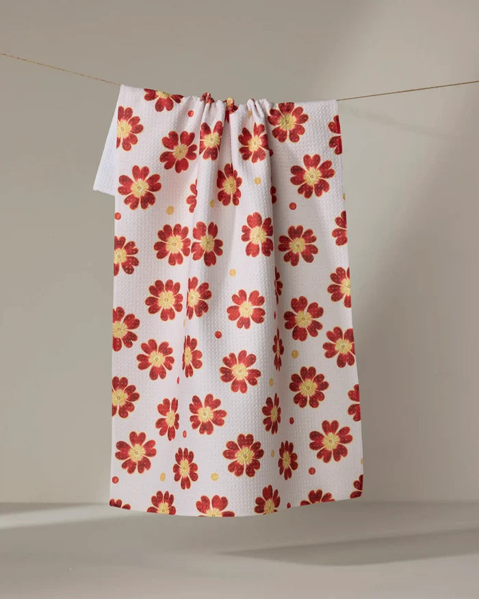 The Spotty Floral tea towel by Linda Fox, crafted from recycled materials, features a white background with red and yellow flowers and hangs neatly over a thin line against a neutral backdrop.