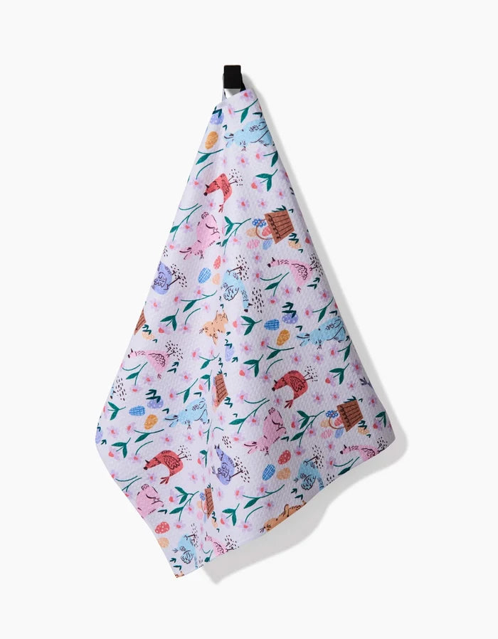 The Spring Bunnies tea towel by Charlie Rowan Designs, made from recycled materials, showcases playful animal, flower, and leaf patterns in pink, blue, orange, and green and hangs from a black loop against a white background.
