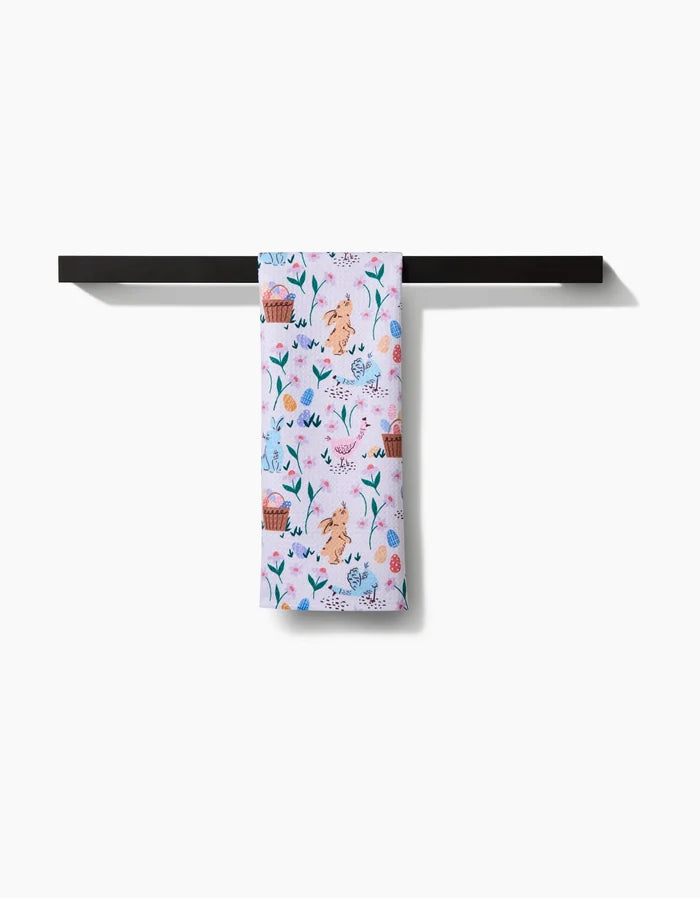 The Spring Bunnies tea towel by Charlie Rowan Designs, made from recycled materials, features a playful animal and nature pattern in light pink and hangs on a sleek black towel bar against a white background.
