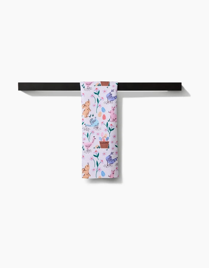 The “Spring Bunnies” towel by Charlie Rowan Designs, featuring pastel hues and whimsical animal and plant prints, hangs neatly on a black horizontal towel bar against a white backdrop.