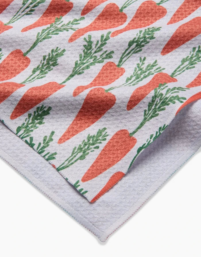 A close-up of the Spring Carrot Hill tea towel by Hufton Studio, showing its white textured fabric with a repeating pattern of orange carrots and green leaves, folded to reveal both the vibrant front and plain underside.