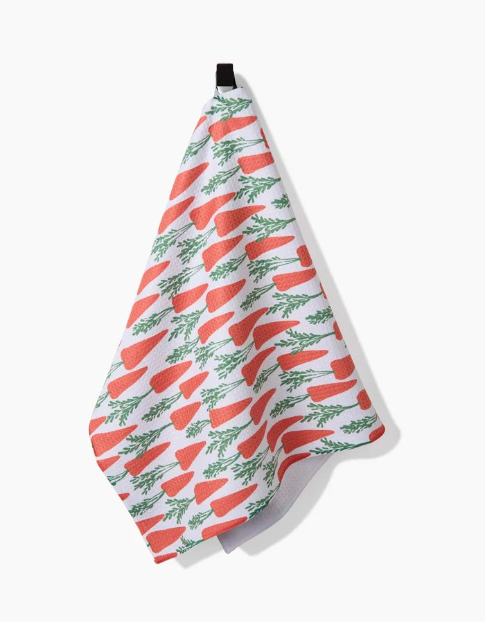 The Spring Carrot Hill kitchen towel by Hufton Studio, made from white recycled fabric, features a repeating orange carrot and green leaf pattern and hangs from a black loop against a plain white background.