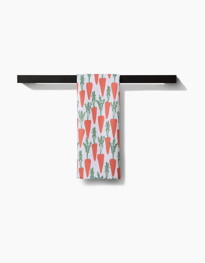 The Spring Carrot Hill tea towel by Hufton Studio, with a red-orange carrot and green top pattern, hangs on a black bar against a white background.