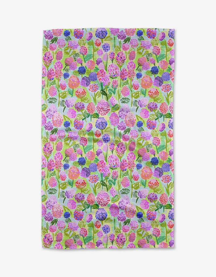 The Elisabeth Wing Spring Clovers tea towel is rectangular, made from soft Geoweave fabric, and features a colorful floral pattern with pink, purple, orange, and blue flowers plus green leaves on a light background.