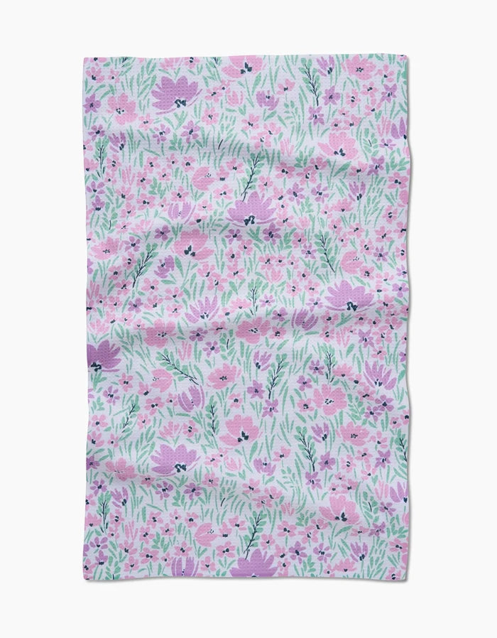 The Hufton Studio Spring Fleur Blooms Geoweave™ tea towel features a floral design with pink and purple flowers and green leaves on a light background, displayed with visible creases and wrinkles.