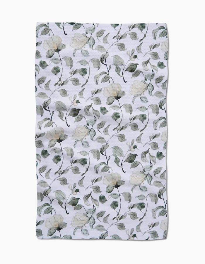 The Spring Floral Blossoms tea towel by Aubrey Fairchild features a white Geoweave fabric adorned with delicate green leaves and cream flowers. Its slightly wrinkled texture enhances the natural charm of this elegant kitchen accessory.