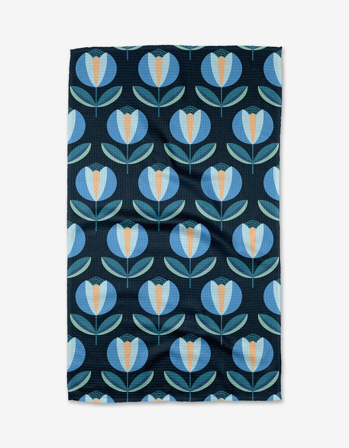 The Blissful Flamingo Spring Florals Tea Towel is made from recycled materials and features a repeating pattern of stylized blue and light blue flowers with orange centers and green leaves on a dark background.
