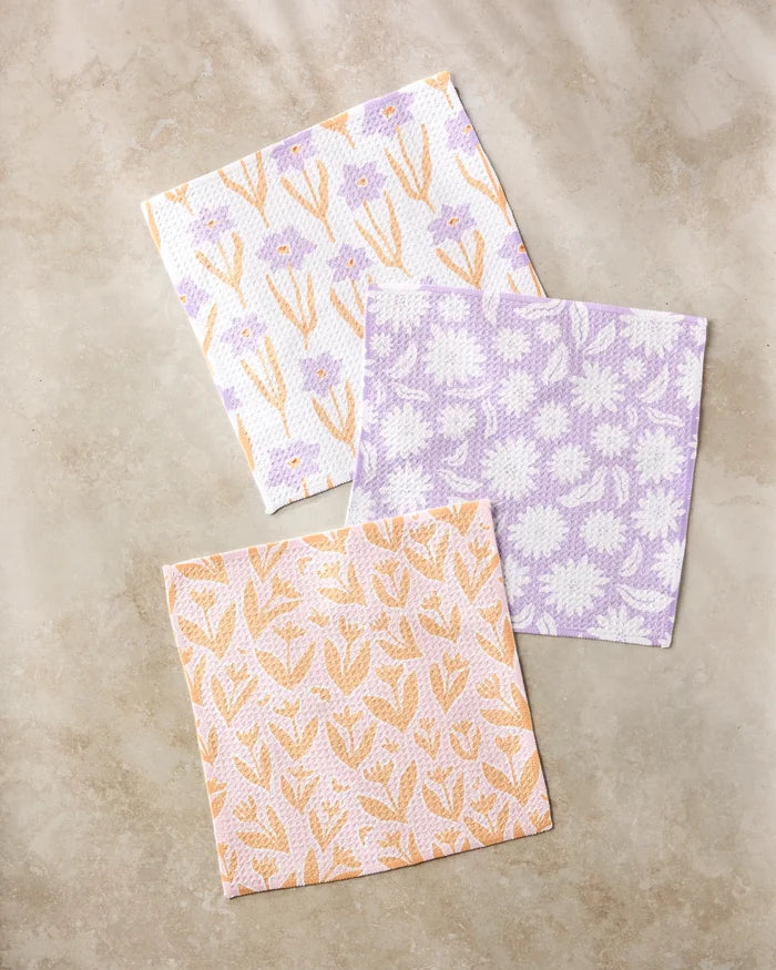 The Kayla Ann Spring Flowers set features three square, super-absorbent dishcloths in lavender, peach, and white, each with a unique floral pattern, beautifully displayed on a beige marble surface.