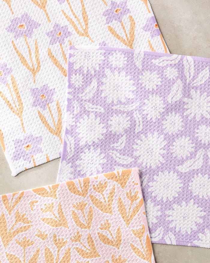 Three overlapping "Spring Flowers" dishcloths by Kayla Ann are shown on a light surface, each super-absorbent white cloth featuring pastel purple and yellow botanical patterns.