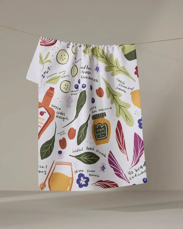 The Spring Harvest Salad tea towel by Janna Sue Design hangs on a line, featuring vibrant illustrations of veggies, fruits, herbs, and recipe notes in multiple colors on a white background.