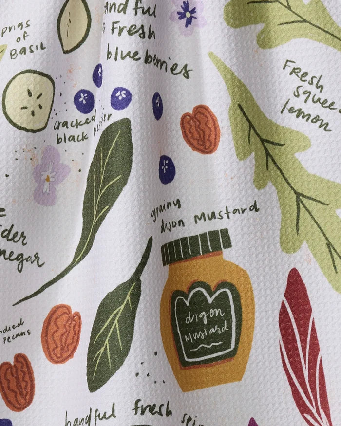 The "Spring Harvest Salad" tea towel by Janna Sue Design features basil leaves, blueberries, lemon, dijon mustard, and pecans in hand-drawn style with handwritten green and black labels on a textured white background.