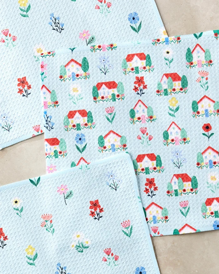 The Ceyda Alasar Spring House Blossom set includes three light blue kitchen towels: two with small colorful flowers and one decorated with rows of red-roofed houses and trees, all displayed on a light background.