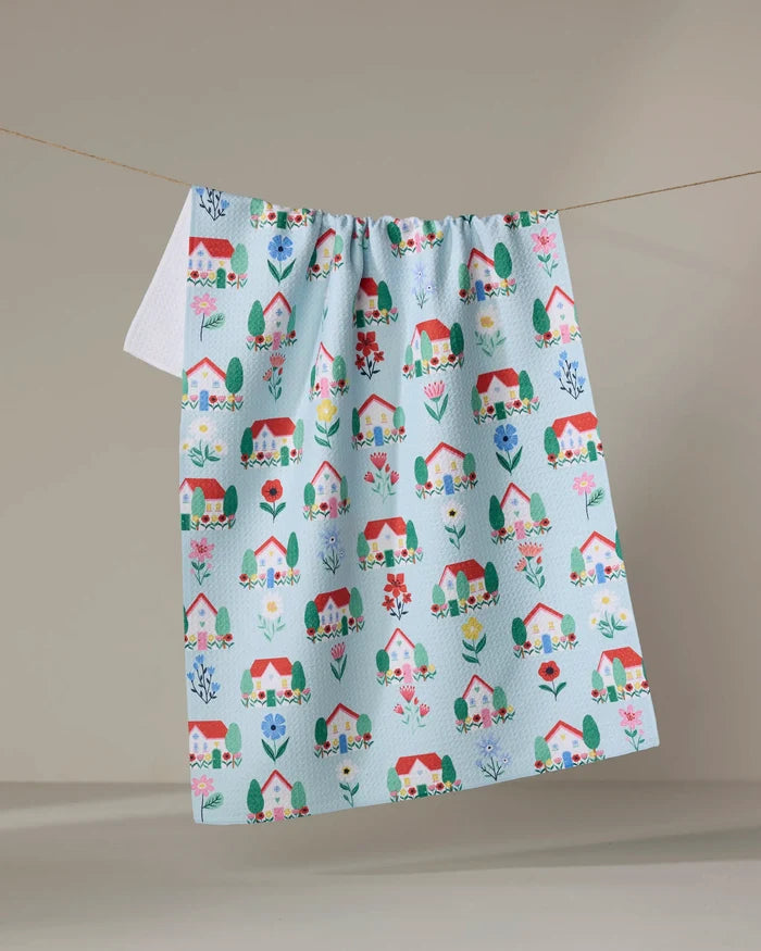 The "Spring Houses" tea towel by Ceyda Alasar features a colorful pattern of houses, trees, and flowers on light blue fabric and hangs on a clothesline against a neutral background.