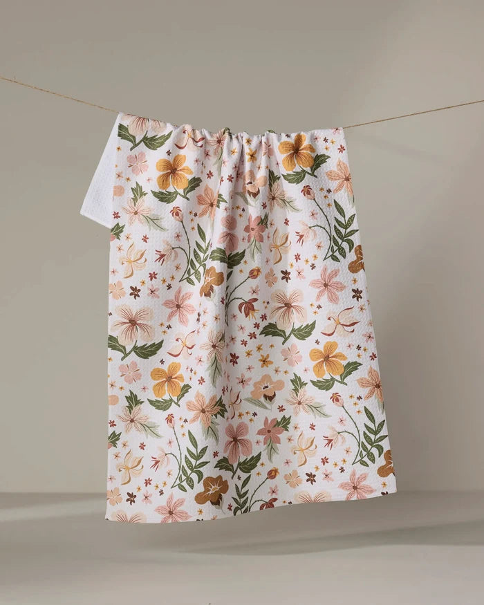 The "Spring In Devon" tea towel by Danelys Sidron features a vibrant floral pattern in orange, pink, and green on white, hanging on a clothesline against a neutral background.