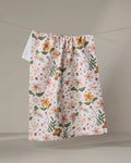The "Spring In Devon" tea towel by Danelys Sidron features a vibrant floral pattern in orange, pink, and green on white, hanging on a clothesline against a neutral background.