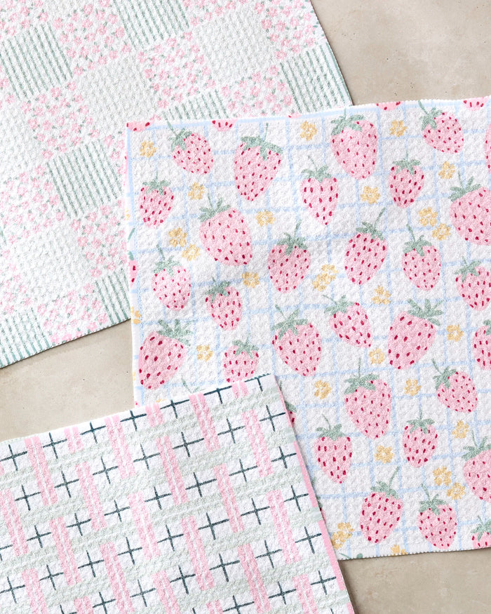 The Spring Picnic Day dishcloth set by Hufton Studio includes three super-absorbent cloths: pink strawberries with yellow flowers, a pink and green plaid, and pink stripes with black X patterns—all made from recycled materials.