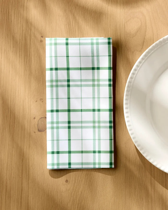 The Hufton Studio Spring Picnic Plaid, a folded white reusable table napkin with green and dark green plaid, rests on a light wooden table beside a white scalloped plate as soft sunlight casts gentle shadows.