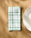 The Hufton Studio Spring Picnic Plaid, a folded white reusable table napkin with green and dark green plaid, rests on a light wooden table beside a white scalloped plate as soft sunlight casts gentle shadows.