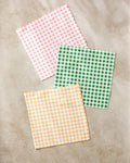 Three Spring Plaid napkins by Danelys Sidron, made from recycled materials, are displayed on a light beige surface. The set includes pink and white, green and white, and yellow and white gingham checkered patterns.