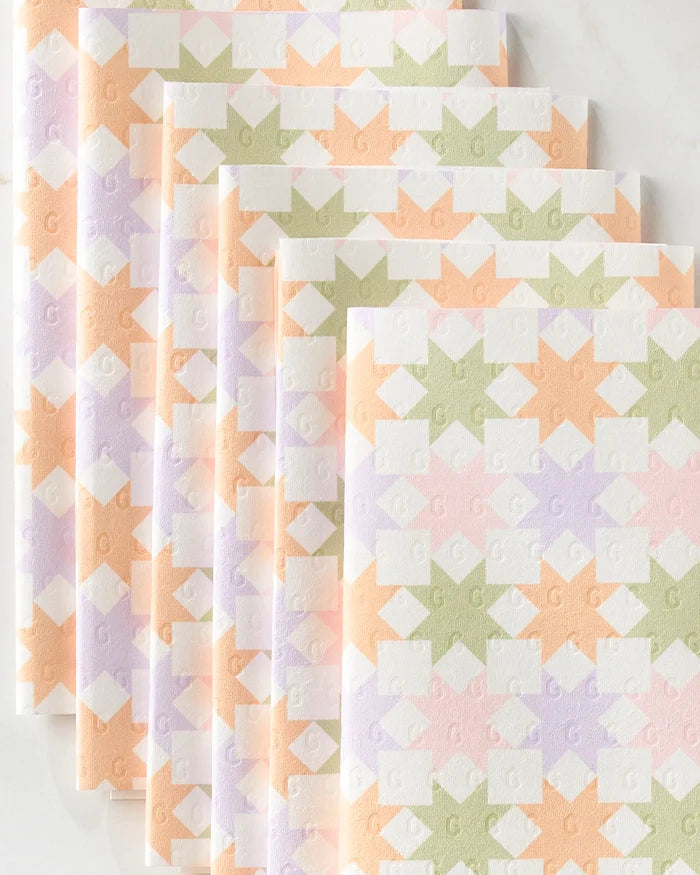 Five folded Kayla Ann Spring Quilt kitchen towels in pastel peach, lavender, green, and white with a geometric diamond and arrow pattern are arranged in an overlapping diagonal stack on a light surface.
