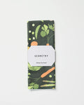 A folded recycled kitchen towel by GEOMETRY, called Spring Sprout, featuring a colorful vegetable pattern with carrots and leafy greens, wrapped in a white label on a plain white background.