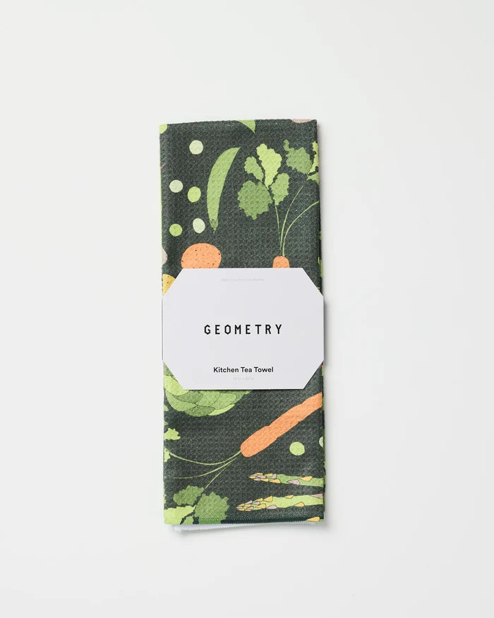 A folded recycled kitchen towel by GEOMETRY, called Spring Sprout, featuring a colorful vegetable pattern with carrots and leafy greens, wrapped in a white label on a plain white background.