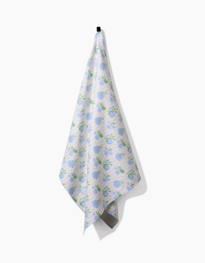 The Spring Wave tea towel by Danelys Sidron, featuring a blue and green floral pattern and made from ultra-absorbent recycled materials, hangs from a black hook against a plain white background.