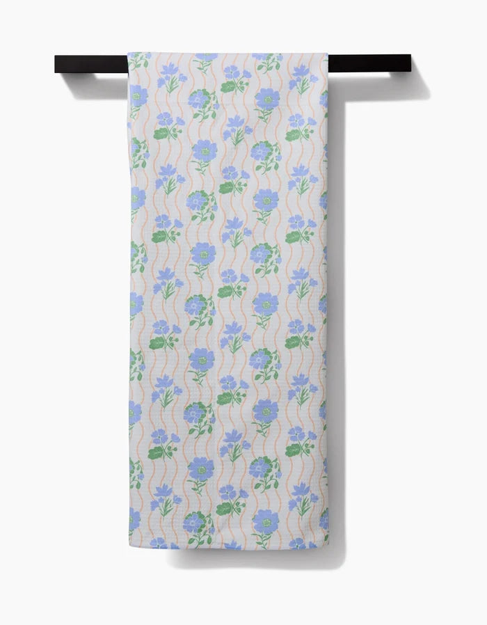 The Danelys Sidron Spring Wave Luxe Bath Towel hangs on a black rod, displaying blue and purple florals with green leaves and peach waves, all on a light background. Made from ultra-absorbent post-consumer recycled materials.