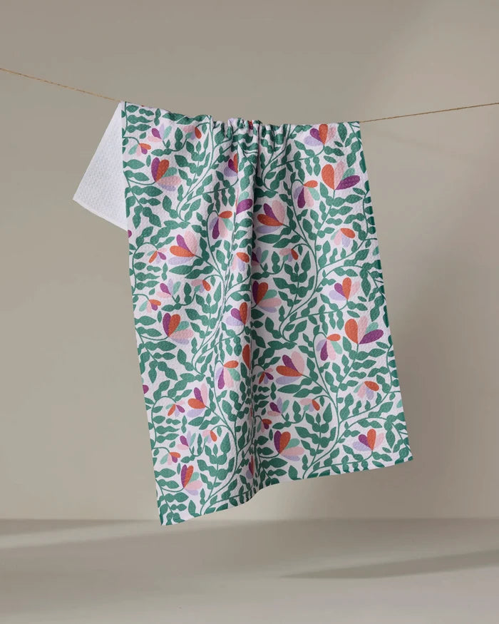 The Claire Iglesias Spring Wavy Leaves Tea Towel, featuring a green leafy and pink floral pattern, hangs on a string against a neutral background—an ultra-absorbent kitchen towel made from recycled materials.