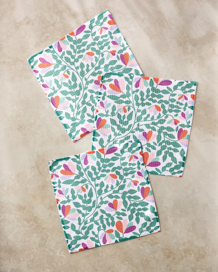Three "Spring Wavy Leaves" square napkins by Claire Iglesias, featuring a vibrant green, orange, and purple leafy floral pattern on recycled fabric, are displayed on a light beige marble surface.