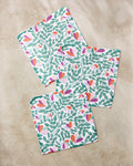 Three "Spring Wavy Leaves" square napkins by Claire Iglesias, featuring a vibrant green, orange, and purple leafy floral pattern on recycled fabric, are displayed on a light beige marble surface.
