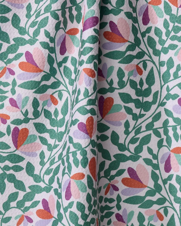 Close-up of a Claire Iglesias Spring Wavy Leaves tea towel, showcasing a vibrant floral pattern with green leafy vines and stylized flowers in pink, purple, orange, and red. The ultra-absorbent towel is textured and gently gathered at the center.