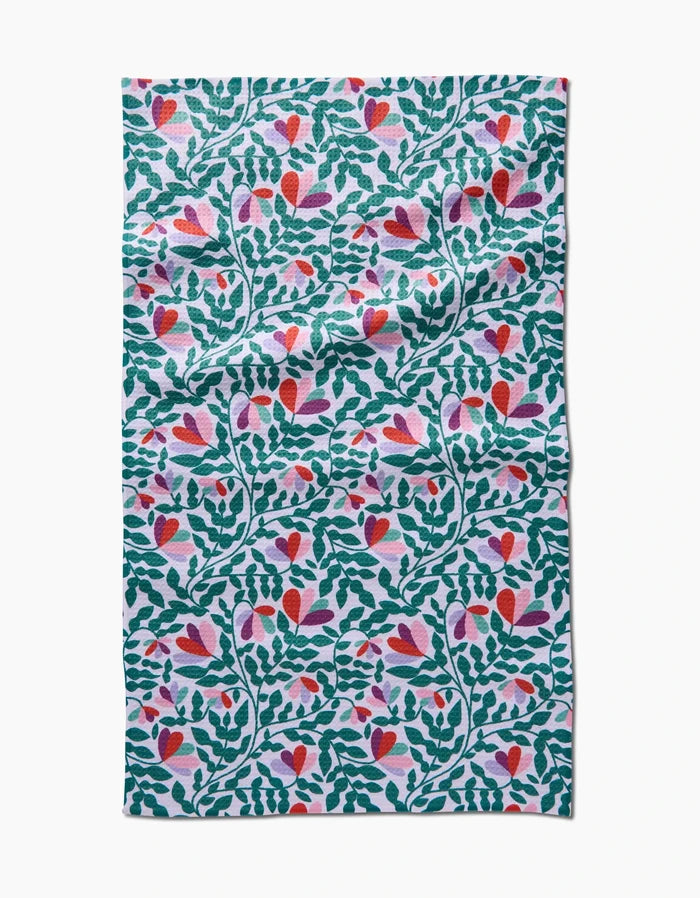 The Spring Wavy Leaves Tea Towel by Claire Iglesias features a vibrant pattern of green leafy branches and pink, red, and purple flowers on white Geoweave fabric. The cloth has a slightly wrinkled texture.