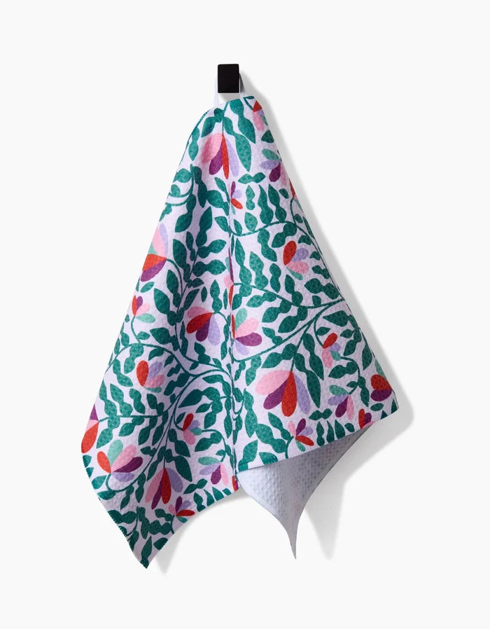 The Spring Wavy Leaves bar towel by Claire Iglesias, made from Geoweave fabric, features a green, red, and pink leafy floral pattern and hangs from a black loop against a white background.