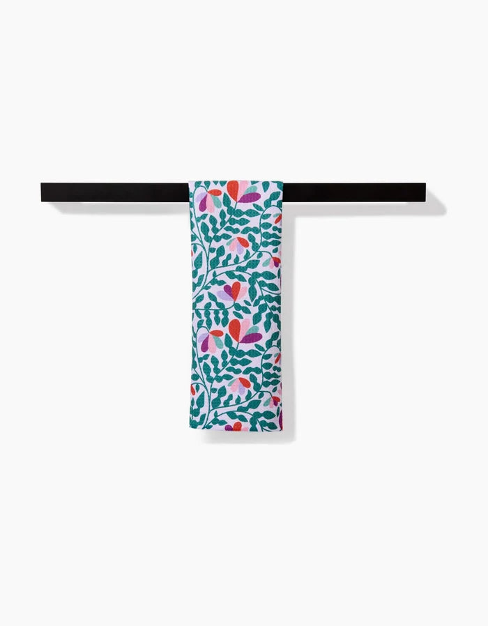 The Claire Iglesias Spring Wavy Leaves bar towel features a vibrant green, red, pink, and purple floral and leaf pattern. Made from soft, durable Geoweave fabric, it’s shown draped over a black bar against a white background.