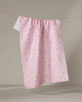 The Blissful Flamingo Springtime Wonders tea towel, made from recycled materials with pink fabric and scattered light red, orange, and yellow teardrop shapes, hangs on a clothesline against a neutral background.