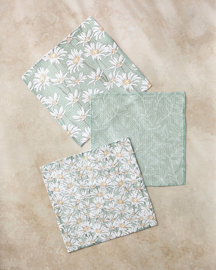 Three "Springtime" fabric napkins by Aubrey Fairchild sit on a beige surface: two display white daisies on light green, while the center napkin has a delicate white leaf pattern, all crafted from recycled kitchen towels.