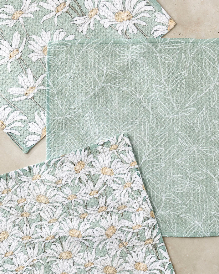 Three textured green cloths from the Aubrey Fairchild "Springtime" recycled kitchen towel set are arranged on a light beige surface—two with daisies and one with white leaves.