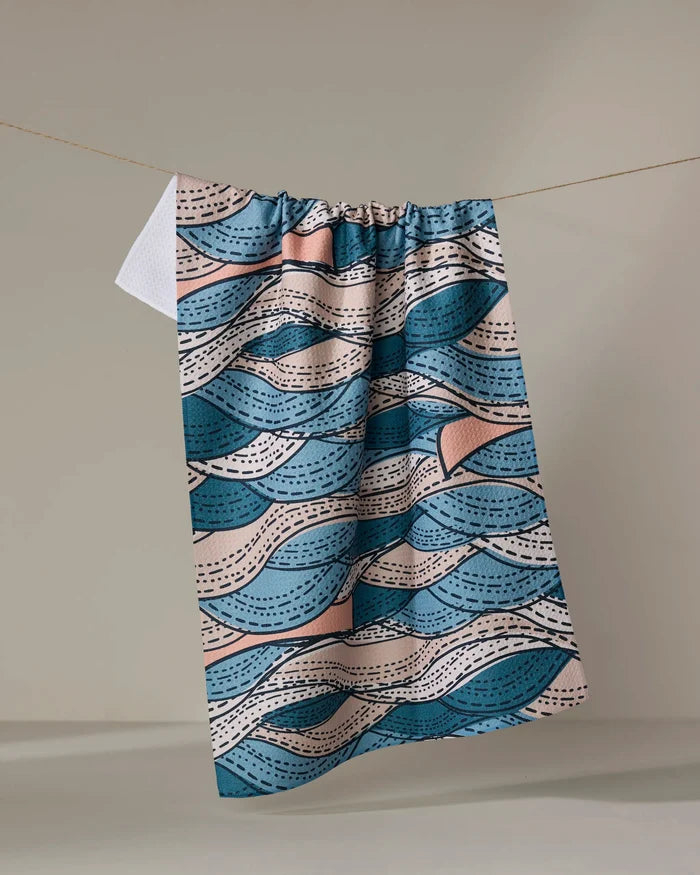 The Twig + Spark “Squall” tea towel, made from Geoweave fabric and featuring blue, pink, and beige wavy patterns, hangs on a clothesline against a plain, light background.