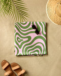 A GEOMETRY Squiggly Micro-loop Beach Blanket with green and pink waves is folded on the sand with black sunglasses on top. Brown sandals, a straw hat, and palm leaf shadows complete this relaxing beach scene.