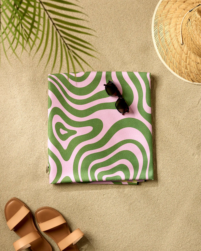 A GEOMETRY Squiggly Micro-loop Beach Blanket with green and pink waves is folded on the sand with black sunglasses on top. Brown sandals, a straw hat, and palm leaf shadows complete this relaxing beach scene.