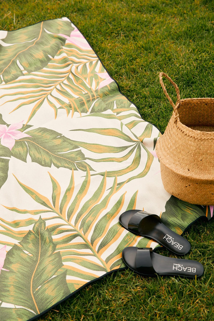 The GEOMETRY Squiggly beach blanket, featuring a tropical leaf pattern and made from quick-drying recycled materials, lies on green grass beside a woven straw basket and black BEACH-labeled slide sandals.
