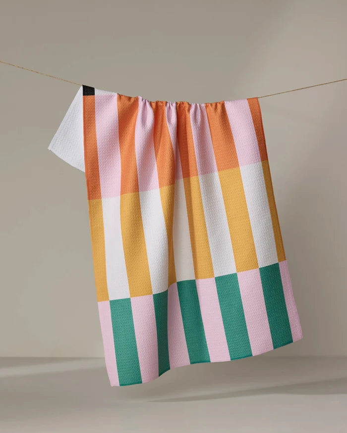 The Megan Roy "Stacked Stripes" tea towel, featuring a colorful checkered pattern with pink, yellow, orange, green, white, and a small black square, hangs on a thin line against a neutral background.
