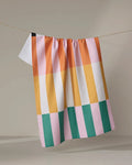 The Megan Roy "Stacked Stripes" tea towel, featuring a colorful checkered pattern with pink, yellow, orange, green, white, and a small black square, hangs on a thin line against a neutral background.