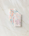 Seven State Flowers notebooks by Julianne Haness, featuring pastel floral designs, are arranged in a fan on a white marble surface next to reusable paper towels, creating an eco-friendly workspace.